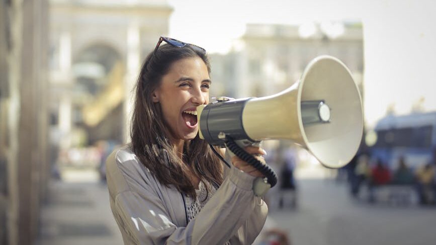 Cheerful young woman in a casual outfit shouting into a megaphone on a sunny day.